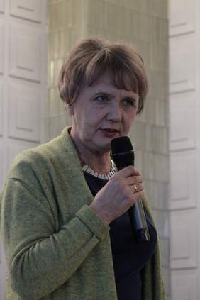 Pokaż zdjęcie: Starsza kobieta z krótkimi włosami w długim zielonym kardiganie stoi z mikrofonem w reku