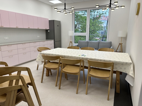 Pokaż zdjęcie: Duży stół 7 osobowy, przy nim drewniane krzesła. Po lewej kuchnia na końcu stoi lodówka. W pomieszczeniu duże okno.