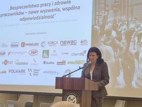 Pokaż zdjęcie: Elegancko ubrana kobieta przemawia do mikrofonu. W tle ekran w logo organizatorów Kongresu.