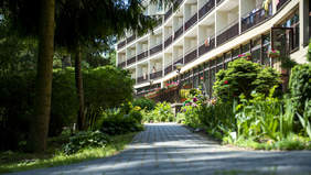 Pokaż zdjęcie: Ośrodek Rehabilitacji Kompleksowej w Nałęczowie. Na zdjęciu alejka otoczona bujną roślinnością, w tle budynek. Na ścianie budynku widać liczne okna i balkony.  