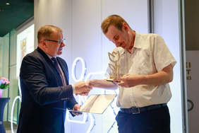 Pokaż zdjęcie: Dwóch mężczyzn elegancko ubranych. Ten z lewej strony wręcza teczkę z dyplomem.