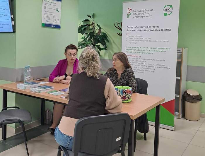 Pokaż zdjęcie: Ekspercki dyżur PFRON w ZUS w Puławach – trzy osoby siedzą przy biurku i rozmawiają; na stole materiały informacyjne, w tle roll-up PFRON i ekran z informacjami.