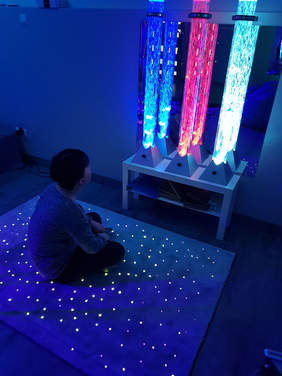 Pokaż zdjęcie: W ciemnym pokoju po turecku, plecami siedzi chłopiec wpatrzony w 3 kolorowe, podświetlone tuby. Tuby wyglądaja jakby był w nich ogień, pierwsza od lewej jest koloru niebieskiego, środkowa czerwonego, po prawej zielonego. Dywan na którym siedzi chł