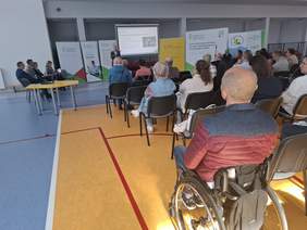 Pokaż zdjęcie: Duża sala. Grupa ludzi siedzi tyłem do widza, obserwując prezentację wyświetlaną na dużym ekranie. Na pierwszym planie widzimy mężczyznę na wózku. W tle prelegent stoi z mikrofonem przed ekranem, obok na krzesłach siedzą pozostali prelegenci.