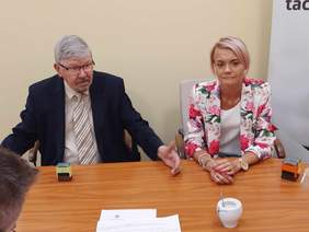 Pokaż zdjęcie: Przy stole siedzi kobieta i mężczyzna. Mężczyzna rozmawia i gestykuluje. Oboje są elegancko ubrani. 