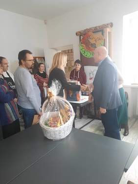 Pokaż zdjęcie: W sali zebrało się kilka osób, na stole stoi kosz z cukierkami, mężczyzna w garniturze przekazuje dyplom i składa gratulacje. Kobieta i mężczyzna wyciągają do siebie ręce