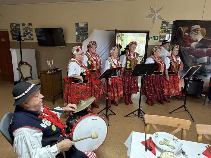 Pokaż zdjęcie: Zbliżenie na zespół ludowy podczas występu. Grupa kobiet w tradycyjnych strojach ludowych śpiewa, stojąc za pulpitami. Na pierwszym planie, z lewej strony, siedzi mężczyzna w ludowym stroju i kapeluszu, grający na bębnie z talerzem.