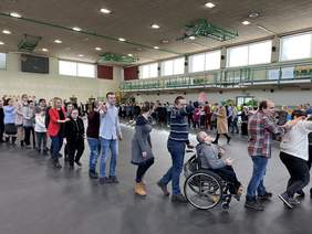 Pokaż zdjęcie: Po szarej podłodze w sali gimnastycznej chodzą soby ustawine w wężka. Na zdjeciu jest też osob ana wózku inwalidzkim. Wszyscy się uśmiechają.
