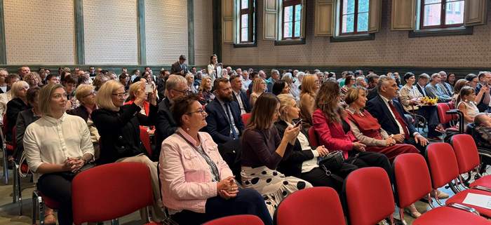 Pokaż zdjęcie: Sala z czerwonymi krzesłami wypełniona publicznością; widoczne są duże okna z drewnianymi ramami i ozdobne ściany. Ludzie siedzą w rzędach, część fotografuje lub korzysta z telefonów, a inni obserwują wydarzenie na scenie.