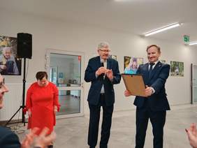 Pokaż zdjęcie: W sali domu mieszkalnego stoi kobieta i dwóch mężczyzn. Jeden z panów przemawia do mikrofonu, a drugi odbiera dyplom z gratulacjami. 