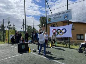 Pokaż zdjęcie: Kobieta ubrana w dżinsy i białą koszulkę robi wymach w tył wyrzucając piłkę. Na siatce otaczającej boisko wisi napis warsztaty terapii zajęciowej igrzysk olimpijskich Chmielno oraz flaga z logo igrzysk olimpijskich. Przed kobietą stoją sędzio