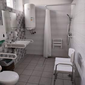 Pokaż zdjęcie: Zdjęcie łazienki, w tle na ścianie wisi piec gazowy, po prawej od piecyka zamontowana jest zasłona prysznicowa. Po lewej stronie od wejścia jest toaleta, umywalka nad którą wisi lustro, zamontowane są pojemniki na mydło i płyn odkażający. Przy toalecie i zlewie znajdują się uchwyty. Po stronie toalety i zlewu na górze jest poziome okno. Na przeciwko zlewu po prawej stronie stoją dwa białe krzesła