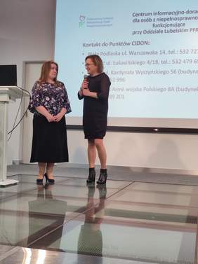 Pokaż zdjęcie: Na scenie stoją dwie kobiety, jedna z nich przedstawia drugą. Panie skierowane są lekko w swoją stronę.