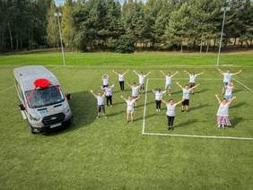 Pokaż zdjęcie: Udekorowany mikrobus zaparkowany jest na boisku otoczonym drzewami. Obok pojazdu znajduje się grupa kilkunastu osób z uniesionymi do góry rękoma.