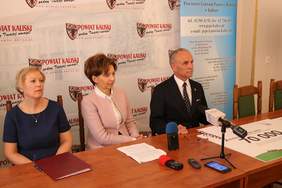 Pokaż zdjęcie: Tzy osoby siedzą przy stole. konferencyjnym, na którym stoi mikrofon i leżż dyktafony dziennikarzy..