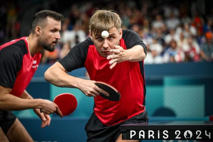 Pokaż zdjęcie: Tenista stołowy z paletką i piłeczką pingpongową w trakcie gry, w tle inny zawodnik