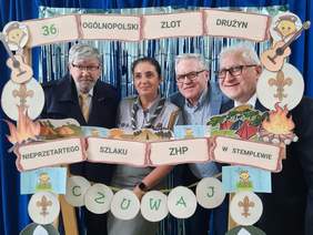 Pokaż zdjęcie: 4 osoby pozujące do zdjęcia. Wśród nich kobieta w harcerskim mundurze. Stoją za okalającą ich ramą z papieru. Widnieje na niej tekst: „36 Ogólnopolski Zlot Drużyn Nieprzetartego Szlaku ZHP w Stemplewie”. 