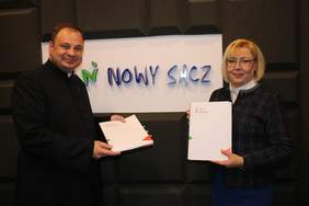 Pokaż zdjęcie: Marta Mordarska – dyrektor Oddziału Małopolskiego PFRON oraz Ks. dr Marcin Kokoszka – ekonom diecezji tarnowskiej - dwie uśmiechnięte osoby stoją obok siebie, trzymają teczki z logotypem PFRON, w papierowych teczkach są umowy dot. projektu