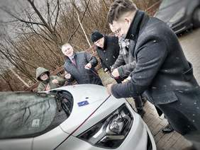 Pokaż zdjęcie: Przy białej toyocie obwiązanej czerwono-niebieskimi wstążkami z nożyczkami stoi kilka osób