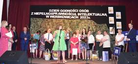 Pokaż zdjęcie: Na scenie w Domu Kultury stoi grupa osób, która przyjmuje i składa życzenia z okazji obchodów dnia godności osób z niepełnosprawnością intelektualną.