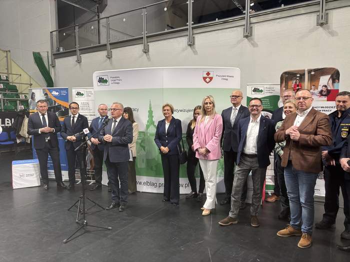 Pokaż zdjęcie: Przy mikrofonie przemawia jeden z uczestników inauguracji wydarzenia, obok stoją przedstawiciele organizatorów oraz zaproszeni goście. W tle widoczne są banery Powiatowego Urzędu Pracy w Elblągu i Miasta Elbląg.