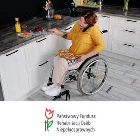 Pokaż zdjęcie: W mieszkaniu w kuchni znajduje sie kobieta siedząca na ręcznym wózku inwalidzkim. Rozmawia przez telefon i przygotowuje jedzenie. Na jej kolanach leży deska z kanapkami. 