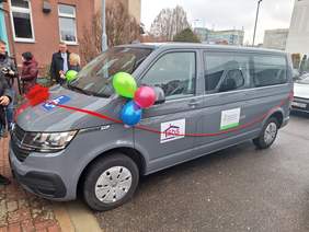Pokaż zdjęcie: Przed budynkiem stoi szary bus. Jest owinięty czerwoną wstążką. Ponadto przybrany jest kolorowymi balonami. Wokół pojazdu stoją ludzie. 