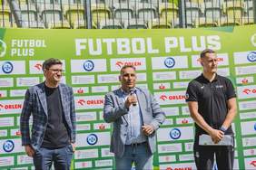 Pokaż zdjęcie: Trzy osoby stoją przed ścianką turnieju Futbol Plus z logotypami sponsorów i partnerów. Osoba pośrodku przemawia do mikrofonu, obok stoją dwaj uczestnicy wydarzenia. W tle widoczne trybuny stadionu.