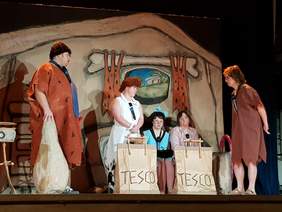 Przedstawienie na scenie. 5 aktorów, sceneria prehistoryczna, najwyższy meżczyzna z lewej stroni podpiera się na wielkiej maczuce