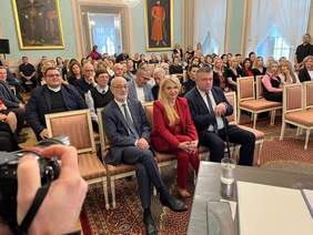 Pokaż zdjęcie: W  sali reprezentacyjnej Trybunału Koronnego zebrali się goście. Goście siedzą na czerwono złotych welurowych krzesłach. Na ścianach wiszą duże obrazy w złotych ramach.