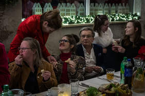 Pokaż zdjęcie: Przy stole siedzą zaproszeni goście, przy jednej z kobiet stanęła kobieta ubrana w czerwony kostium. Kobiety trzymają w ręku kawałek chleba.