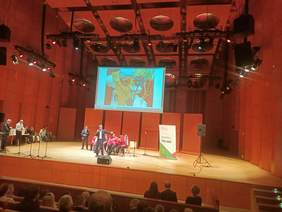 Pokaż zdjęcie: scena sala koncertowa na której występują wokaliści studia integracji. Chłopak jako solista trzyma mikrofon i śpiewa 