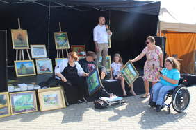 Pokaż zdjęcie: Zdjęcie sceny przy której porozstawiane są obrazy, na scenie siedzi malarz i próbuje sięgnąć stopami po obraz, obok siedzi jego żona, a przed sceną stoi niewielka publiczność