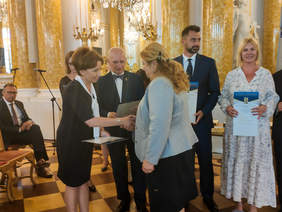 Pokaż zdjęcie: Pani Prezes podaję rękę laureatce konkursu, w drugiej ręce trzyma dyplom. W tle stoja osoby wyróżnione