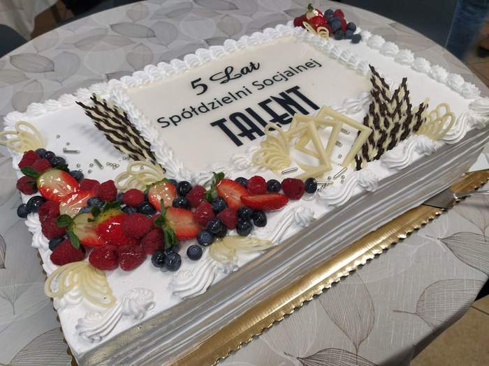 Pokaż zdjęcie: Na okrągłym stoliku stoi duży tort w kolorze białym udekorowany zarówno malinami, truskawkami oraz borówkami, jak i motylami oraz baziami wykonanymi z czekolady. Wewnątrz tortu widnieje napis: „5 lat Spółdzielni Socjalnej Talent”.