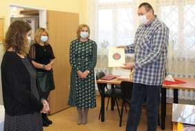 Pokaż zdjęcie: Cztery osoby, trzy kobiety i jeden mężczyzna.  Jedna z nich wręcza mężczyźnie dyplom na którym widzimy czerwone serce na niebieskim kółku.