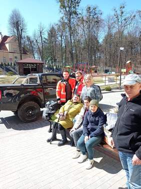 Pokaż zdjęcie: Trzy kobieta siedzą na ławce, obok ławki stoją jeszcze cztery osoby, z tyłu stoi duży czarny samochód, słoneczna pogoda