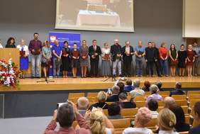 Pokaż zdjęcie: 18 osób na scenie i osoby na widowni, które robią zdjęcia