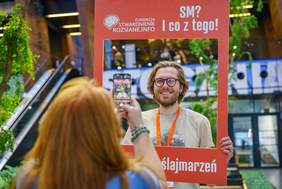 Pokaż zdjęcie: Kobieta robi zdjęcie telefonem mężczyźnie w brodzie i okularach, który trzyma czerwoną ramkę z napisem SM I co z tego! Oraz z logo organizatora.