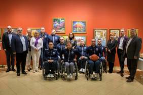 Pokaż zdjęcie: Grupa osób pozyująca na tle obrazów. Wśród zgomadzonych kobiet i mężczyzn sa osoby z niepełnosprawnościami, w tym osoby na wózkach inwalidzkich.