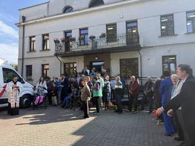 Pokaż zdjęcie: Przed wejściem do budynku stoją podopieczni PSONI w Zamościu oraz Ksiądz i przedstawiciele organizacji w oczekiwaniu na uroczyste poświęcenie nowo zakupionego busa.