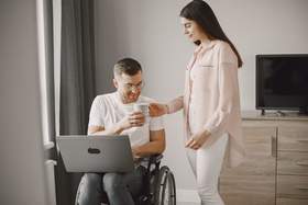 Mężczyzna siedzi na wózku inwalidzkim i trzyma na kolanach laptopa. Obok niego stoi kobieta i podaje mu kubek. W tle widać meble i telewizor. 