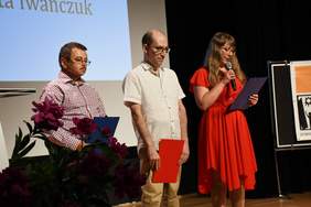 Pokaż zdjęcie: Na scenie stoją 3 osoby dwóch mężczyzn i kobieta, trzymają przed sobą kartki i czytają fragmenty wiersza.