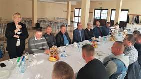 Pokaż zdjęcie: Kobieta stoi przy stole, przemawia. Słuchacze siedzą przy długim stole..