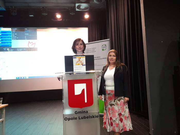 Pokaż zdjęcie: Przy mównicy stoją dwie kobiety, pozują do zdjęcia, w tle baner z logo CIDON.