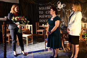 Pokaż zdjęcie: Na scenie stoją trzy kobiety. Blondynka w granatowym garniturze trzyma kwiaty, obok stoi kobieta w ciemnej sukience i kolejna w jasnej marynarce. Kobiety patrzą na siebie i składają sobie życzenia.