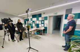 Pokaż zdjęcie: W pomieszczeniu po prawej stronie stoi mężczyzna. Po lewej stronie znajduje się dziennikarka, operator, kamera i statyw. 