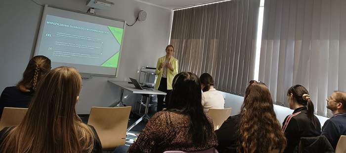 Pokaż zdjęcie: Grupa siedmiu osób siedzi w sali podczas wystąpienia kobiety, która stoi obok okna zasłoniętego żaluzjami. Na dużym ekranie wyświetlone są informacje o Wypożyczalni technologii wspomagających. Obok kobiety znajduje się stolik, a na nim laptop.