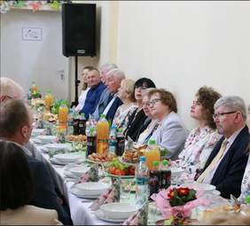 Pokaż zdjęcie: Przy podłużnym stole siedzą ludzie. Są to goście zaproszeni na spotkanie wielkanocne. Na stole znajduje się wiele światecznych potrwa i wielkanocnych dekoracji. 