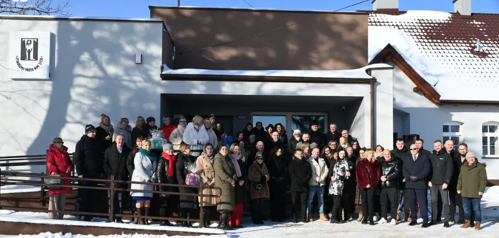 Pokaż zdjęcie: Grupa kilkudziesięciu osób w zimowych płaszczach stoi przed wejściem do jasnego budynku z podjazdem i barierką. Słoneczny dzień, śnieg na ziemi i dachu, nad drzwiami zadaszenie, po lewej stronie znak z symbolem na elewacji.